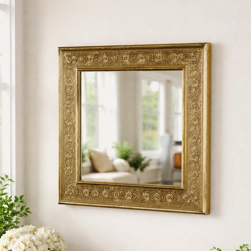 Decorative gold-framed mirror on a white wall with a white cabinet below containing books, a candle, and plants.